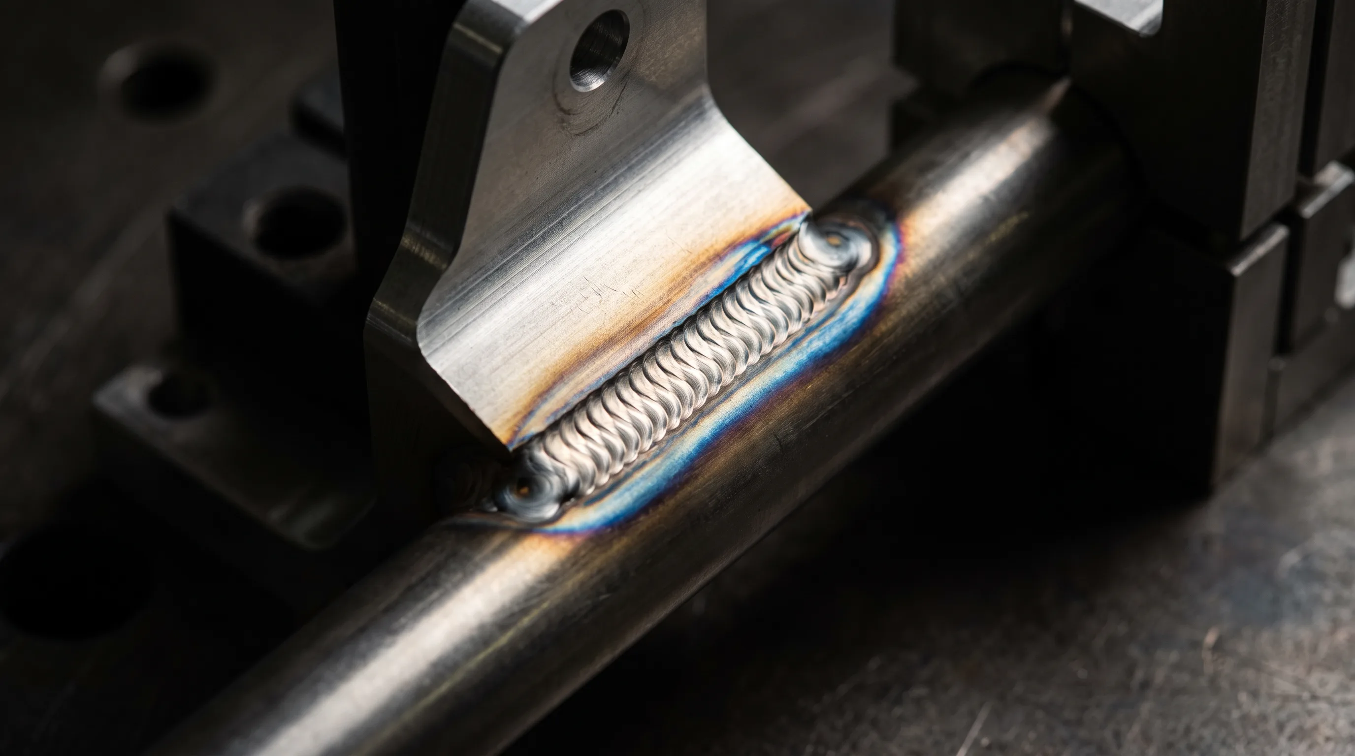 A machined bracket TIG-welded to a steel tube in a fixture: the bead is evenly stacked with clean tie-in at both toes and straw-to-blue heat tint fanning out, the air completely clear.