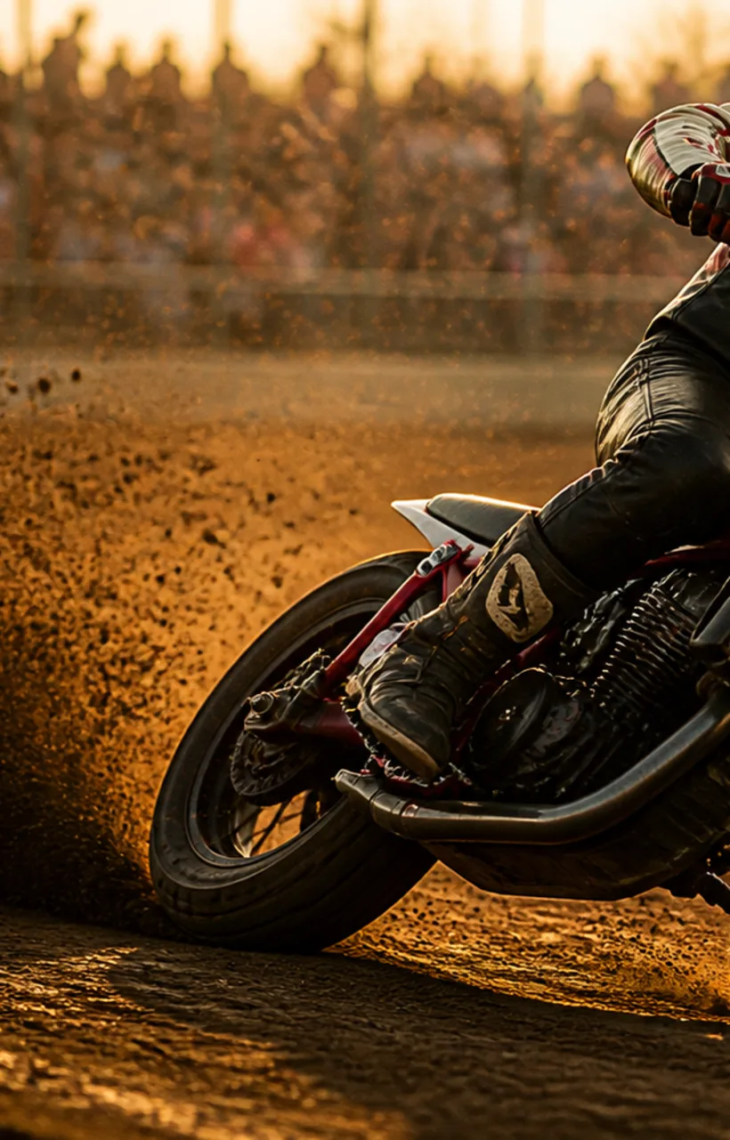 A flat-track race motorcycle sliding through a dirt corner, throwing a rooster-tail of dirt.
