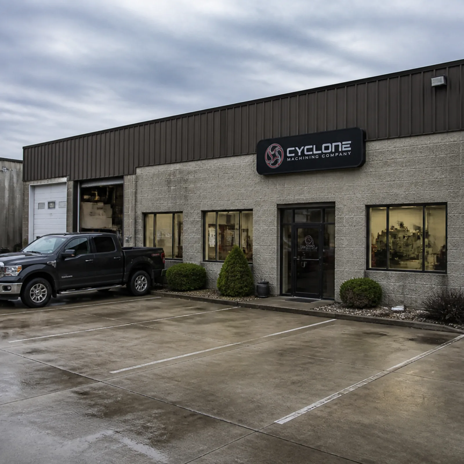 A block-and-steel industrial building under a grey sky, a pickup parked on the wet paving out front.