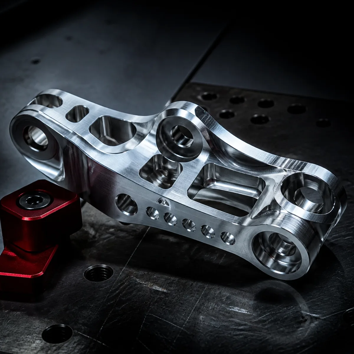 A billet linkage bracket with counterbored pivot bores, lightening pockets and a row of drilled holes along its lower web, lying on a scarred tooling plate beside a red clamp block.