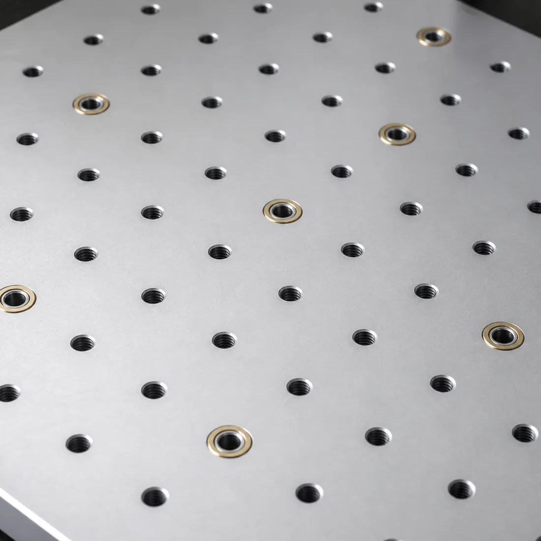 An angled close-up of a machined aluminum modular tooling plate: a dense grid of tapped mounting holes with brass-ringed locating bushings set among them.