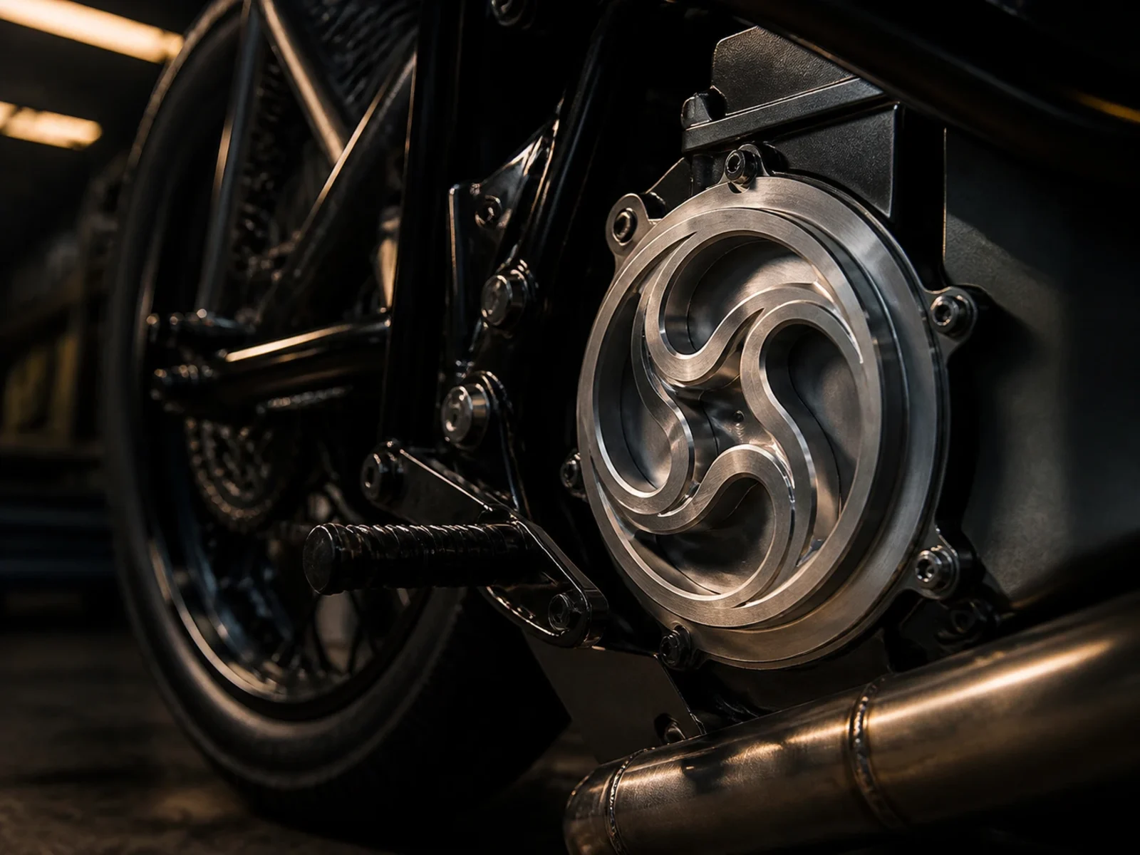 A billet engine cover with a three-lobe swirl cut in deep relief, bolted to the side of a motorcycle, chain and rear wheel visible behind it.